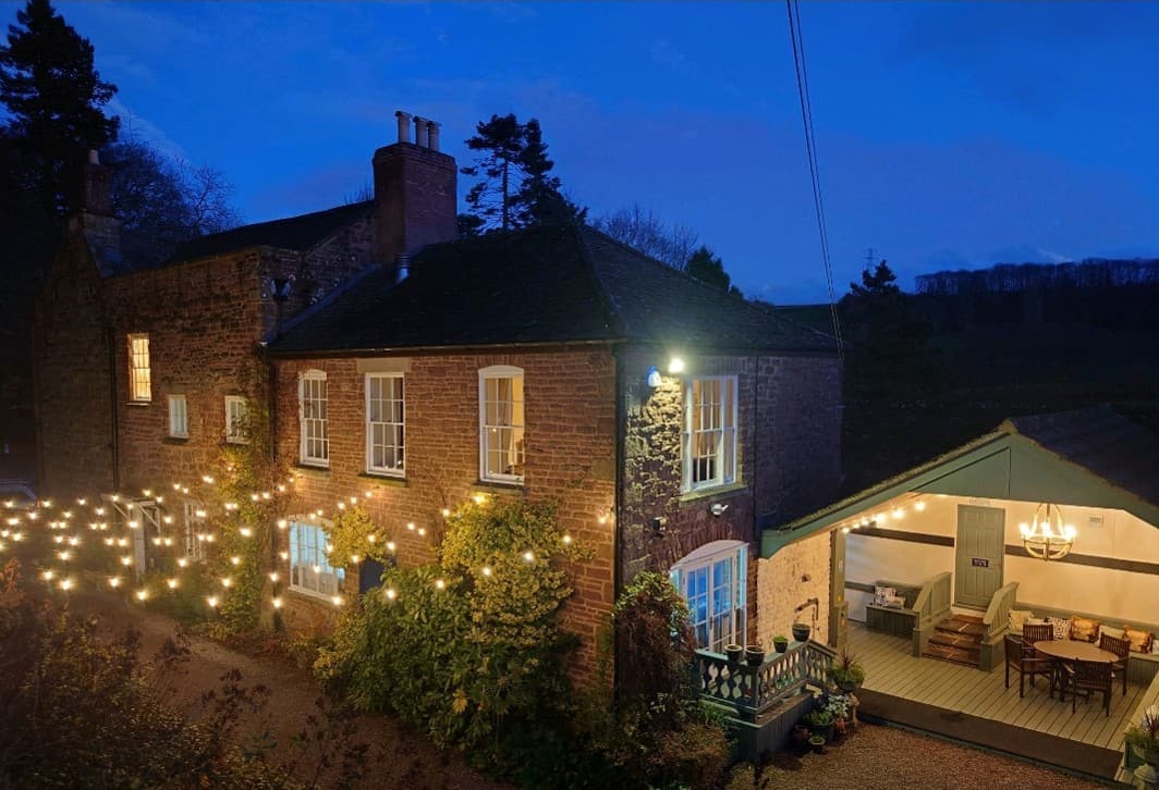 Brick cottage at dusk with holiday lights, illuminated extension, and evening ambiance