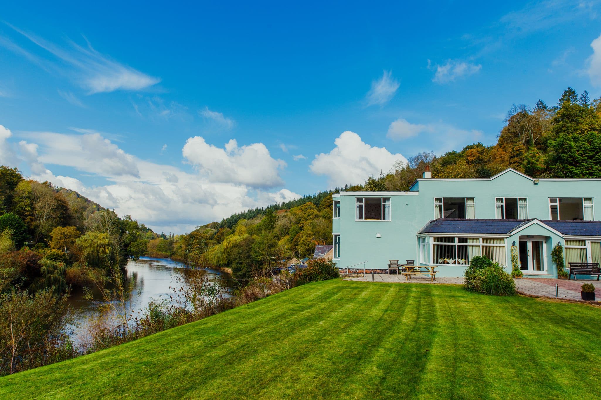 Modern turquoise house with riverside location, manicured lawn and mountain woodland backdrop