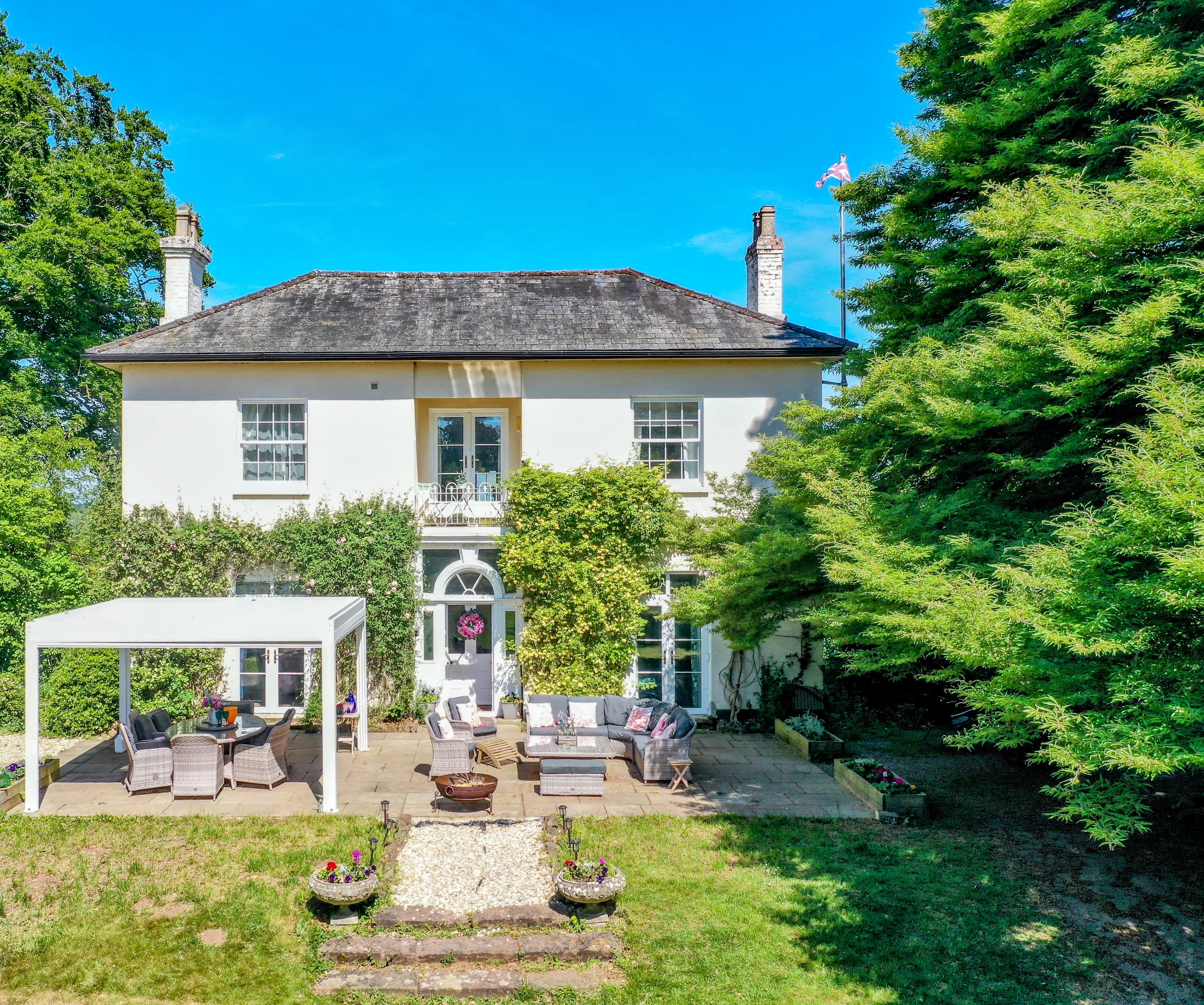 White two-storey country house with ivy, slate roof, patio area and manicured gardens under clear sky