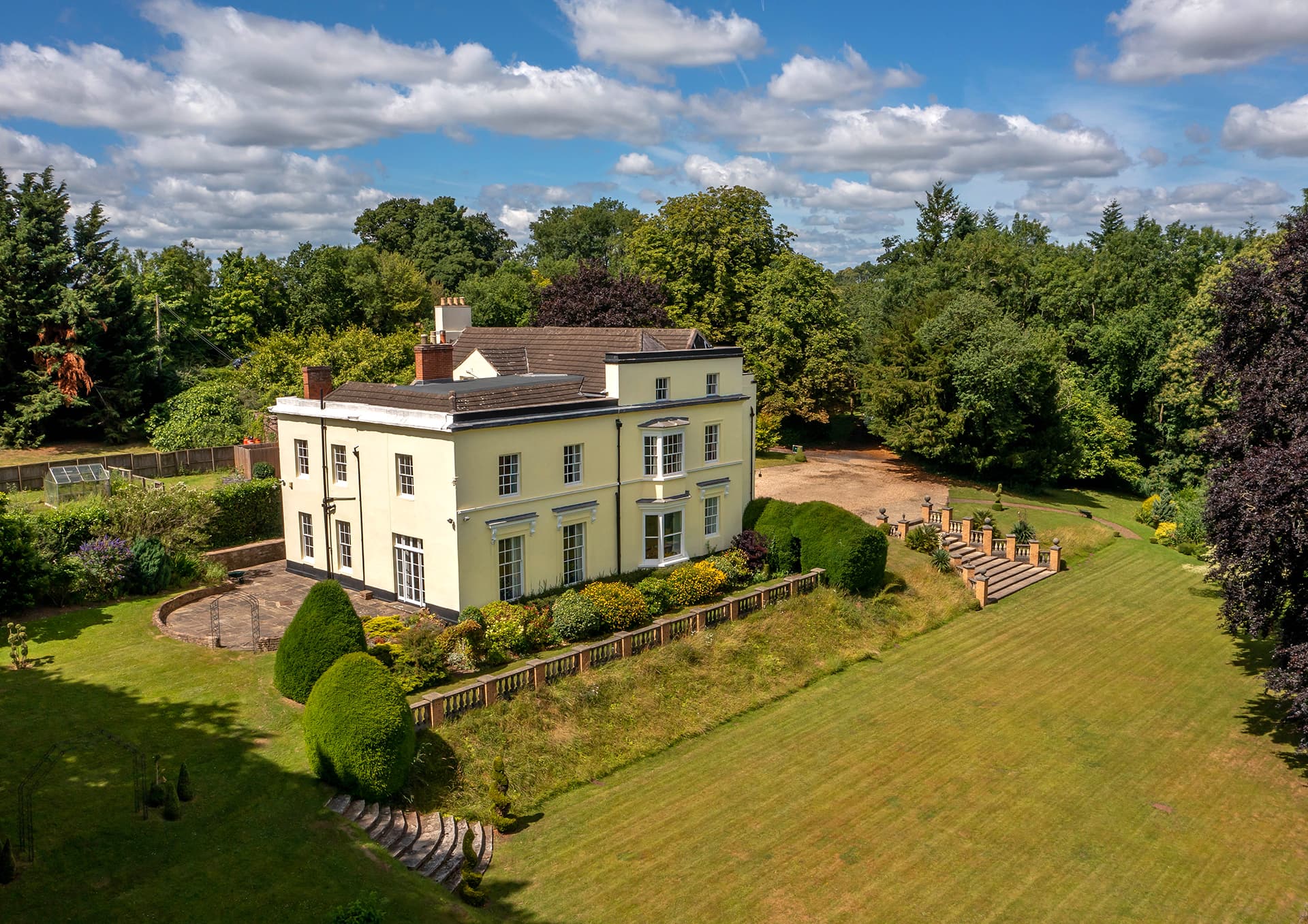 Aerial view of cream and pale yellow country mansion surrounded by manicured lawns and woodland.