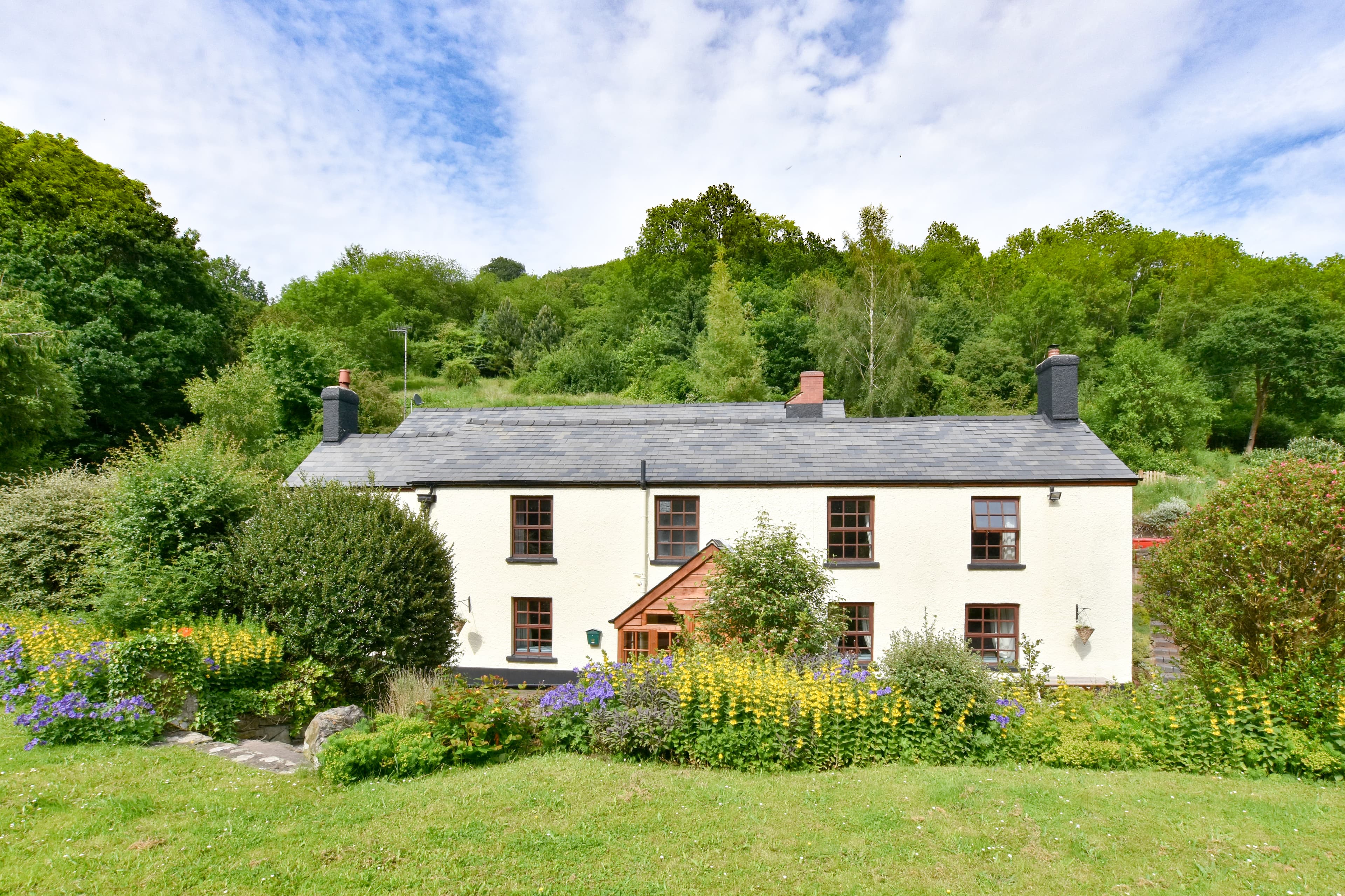 Charming white stone cottage with slate roof, flowering gardens, and natural woodland backdrop.