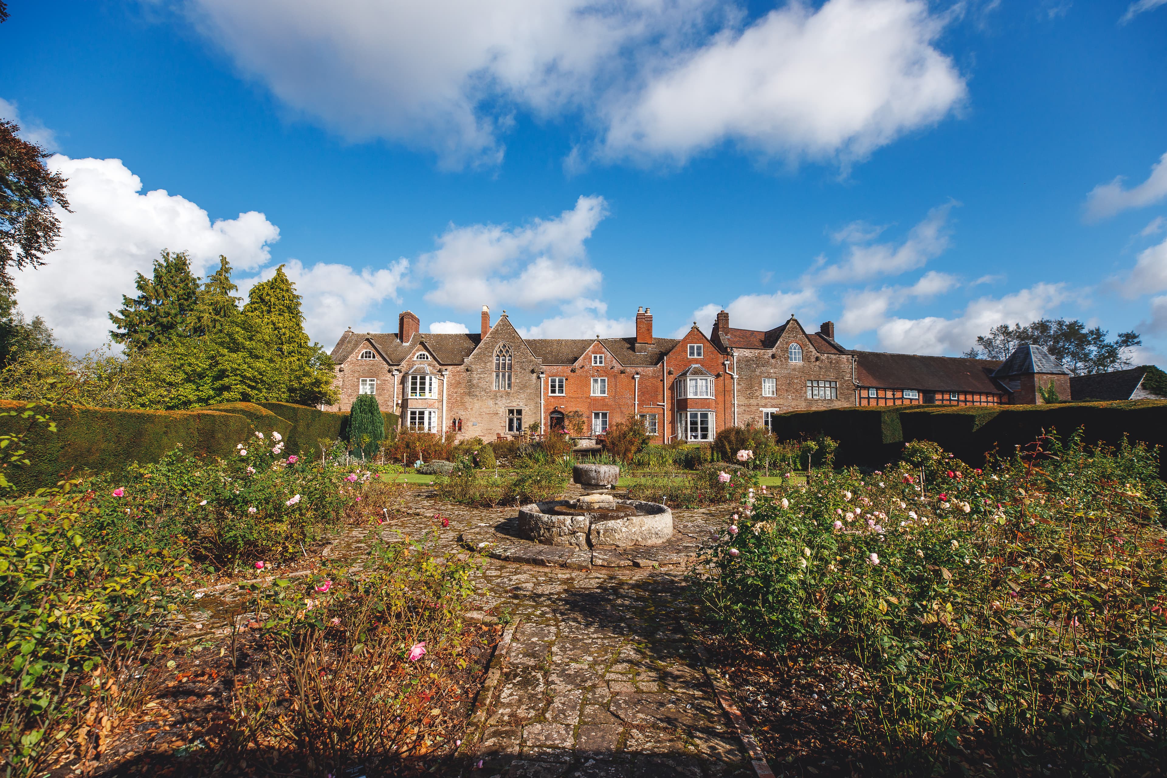 Large period country house with red brick and stone facade, manicured gardens with roses and well-maintained hedges